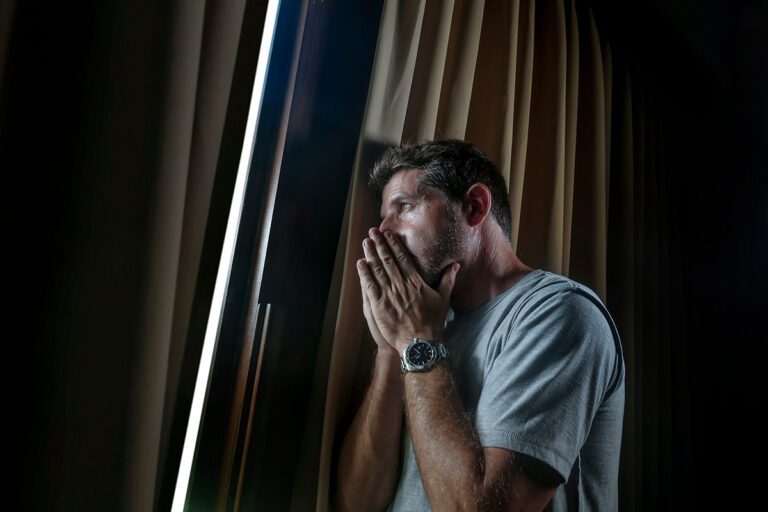 Man inside dark room looking through curtains while struggling with addiction