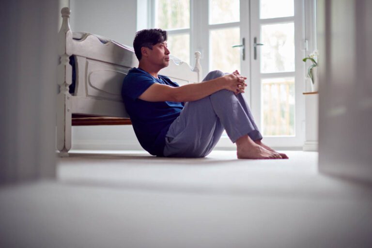 Depressed man sits on floor against bed frame